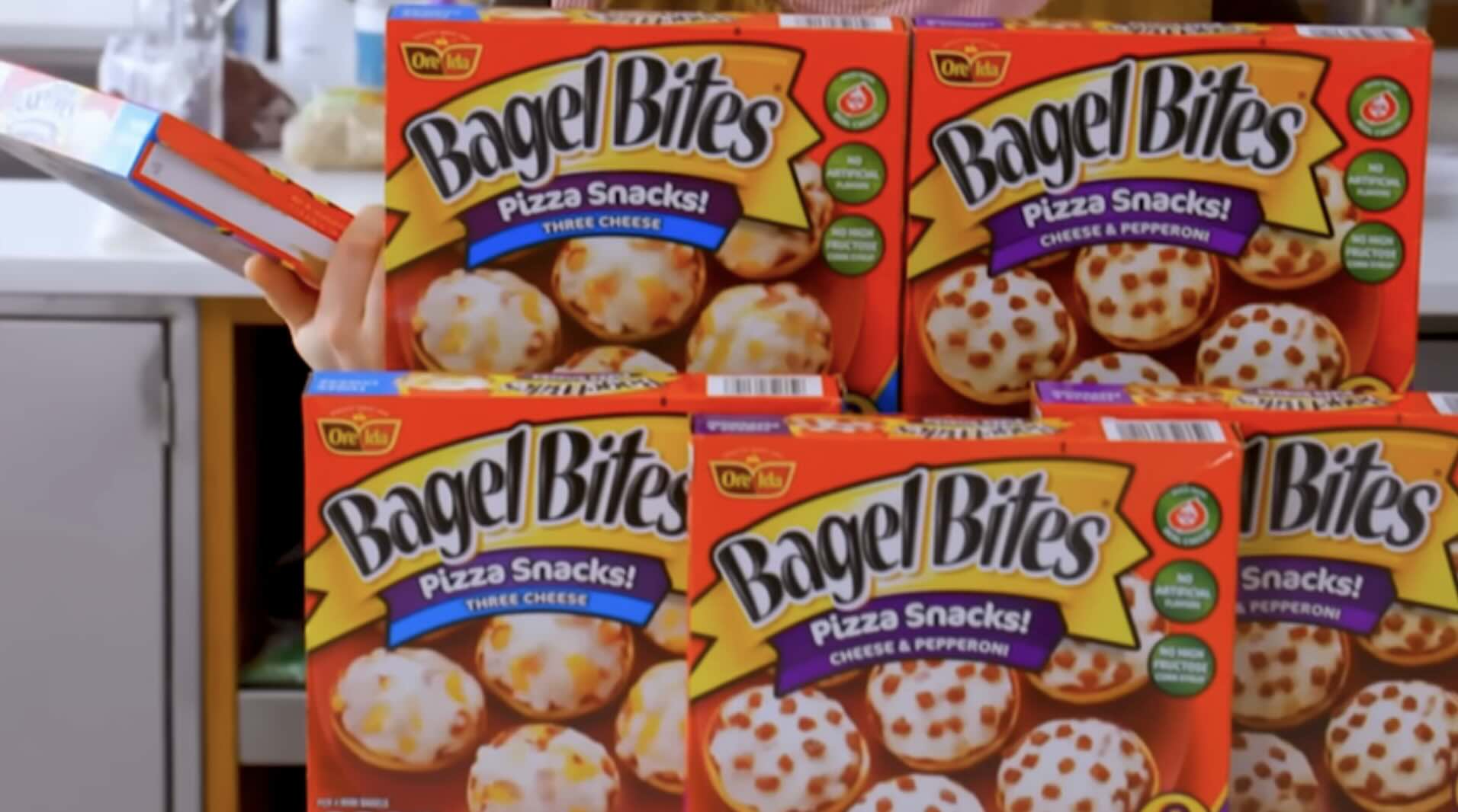 Four boxes of Bagel Bites pizza snacks are stacked on a kitchen counter. Two boxes are three cheese flavor, and two are cheese and pepperoni flavor. A hand is holding one of the boxes on the left side.