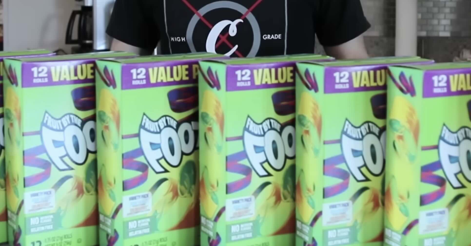 Several brightly colored boxes of Fruit by the Foot snacks labeled "12 VALUE ROLLS" are lined up in front of a person wearing a black shirt with a circular design.