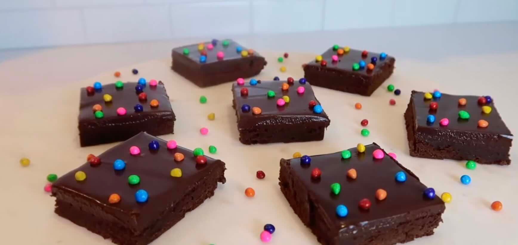 Seven chocolate brownies topped with glossy chocolate glaze and colorful round sprinkles are arranged on a white surface, with more sprinkles scattered around them.