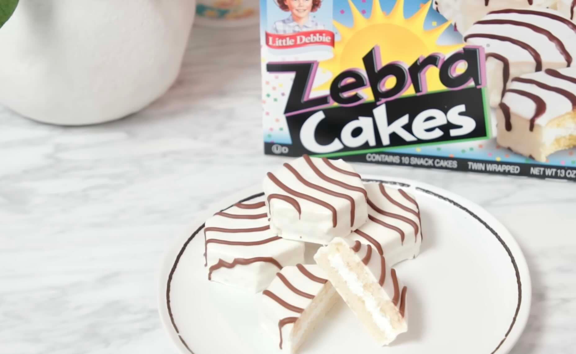 A plate with three Little Debbie Zebra Cakes, one cut in half to show the cream filling, sits in front of a Zebra Cakes box on a white marble surface.