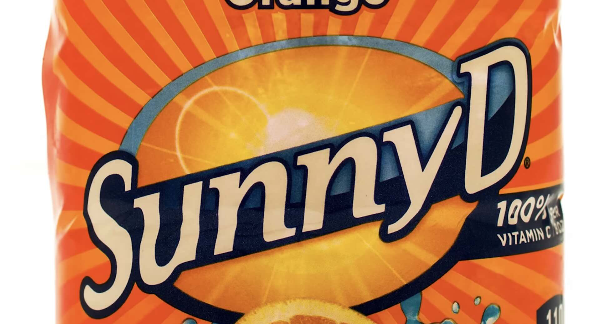Close-up of a SunnyD orange drink label, featuring large, bold "SunnyD" text with an orange slice and rays of light in the background. The design is bright orange and yellow with "100% Vitamin C" also visible.