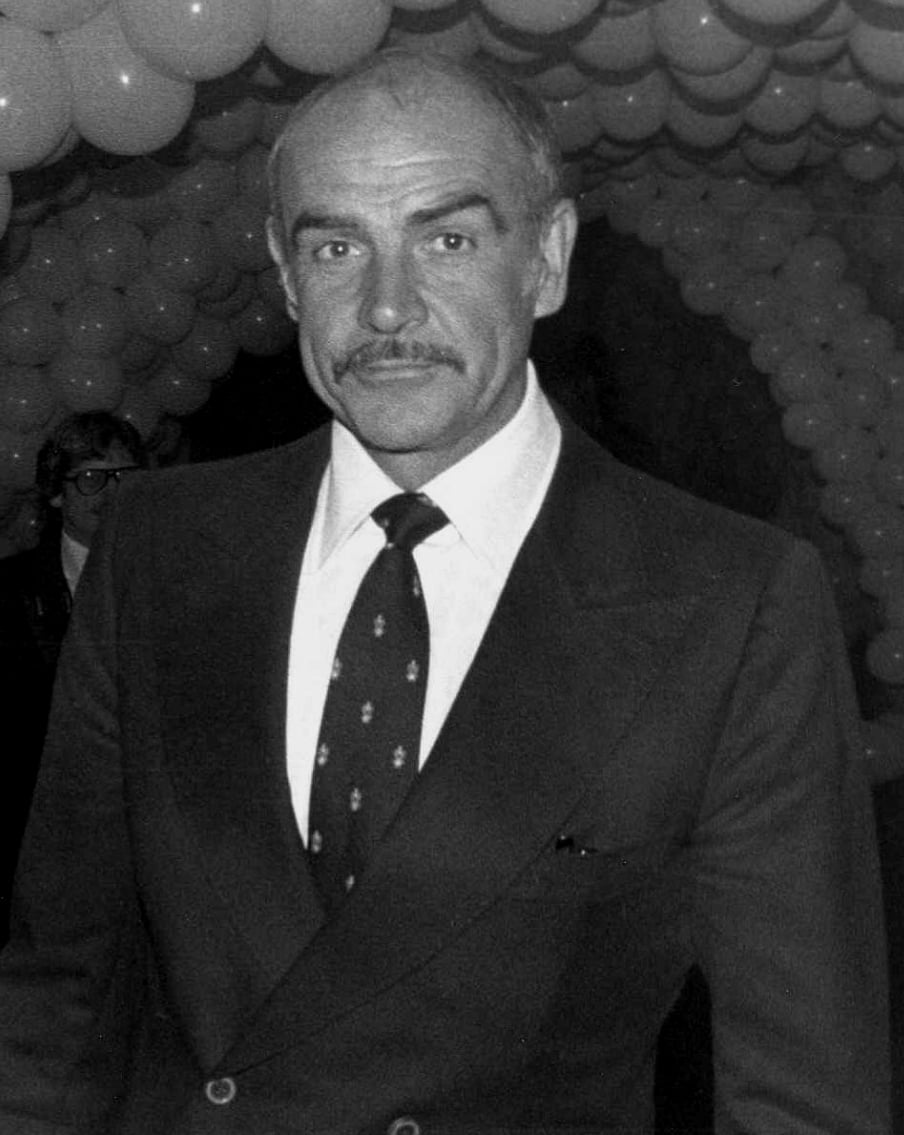 A man in a suit and tie stands in front of an archway made of balloons. He has a mustache, short hair, and is looking at the camera. People and more balloons are visible in the background.