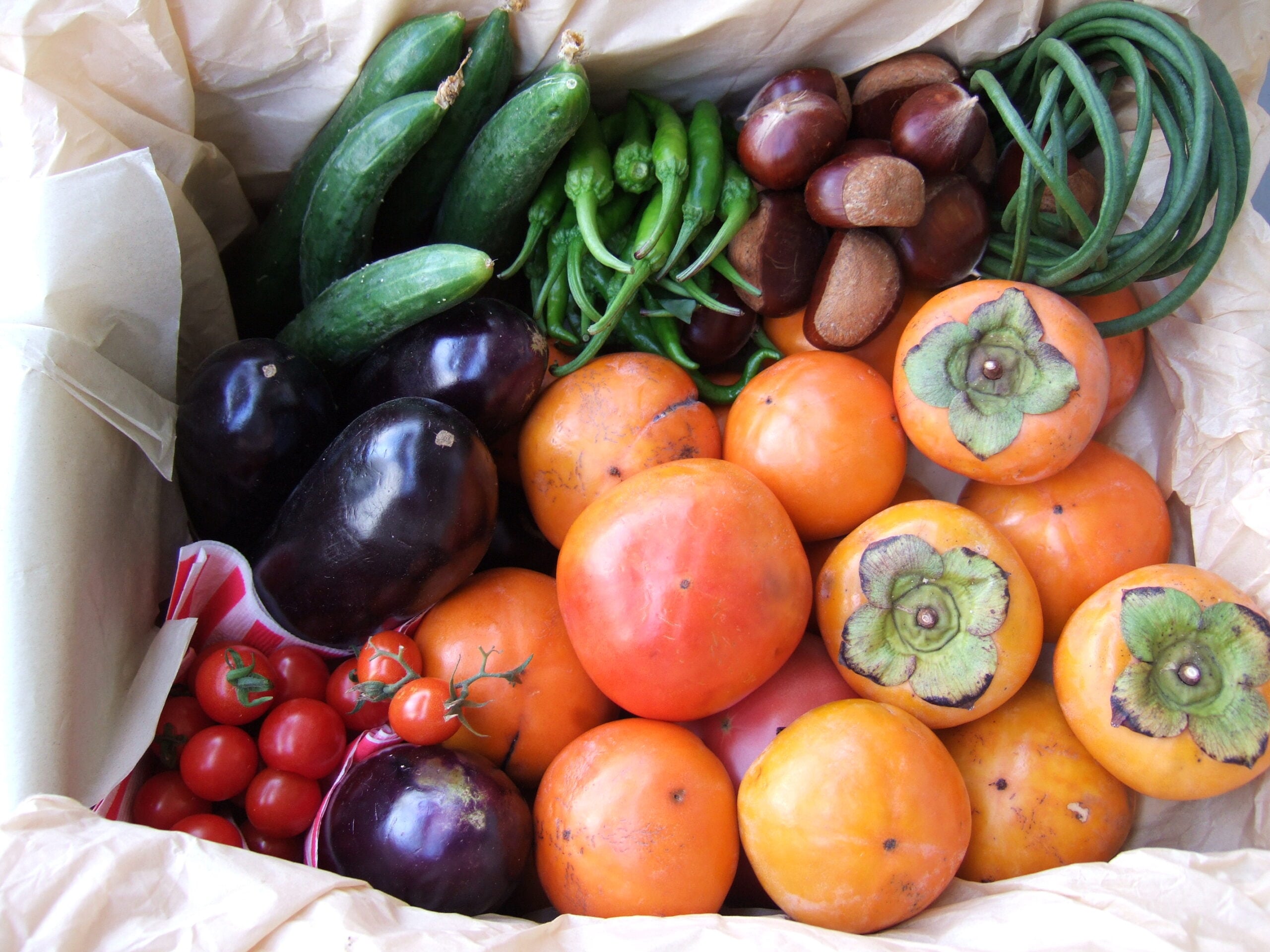 A box filled with assorted fresh produce, including tomatoes, eggplants, cucumbers, green peppers, chestnuts, long green beans, and several orange persimmons.