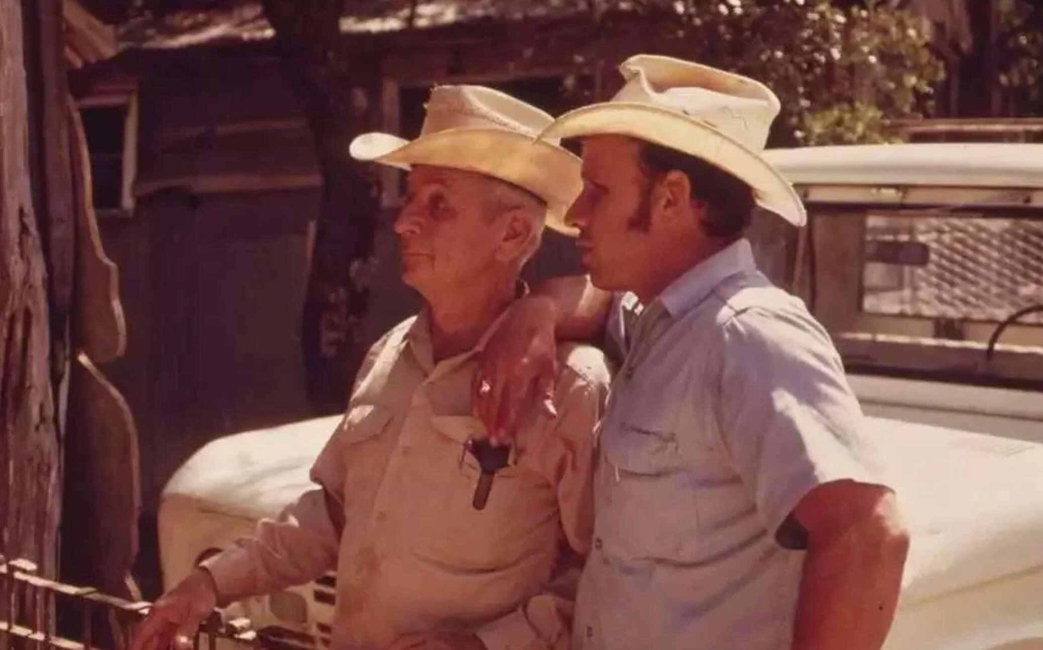 Two men in cowboy hats stand side by side outdoors, one with his arm resting on the other's shoulder. Both wear light shirts and look into the distance, with a white vehicle and wooden fence in the background.
