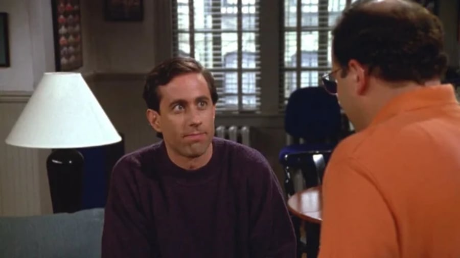 A man in a dark sweater sits on a couch looking at another man in an orange shirt, who is standing with his back to the camera in a room with a lamp and window in the background.