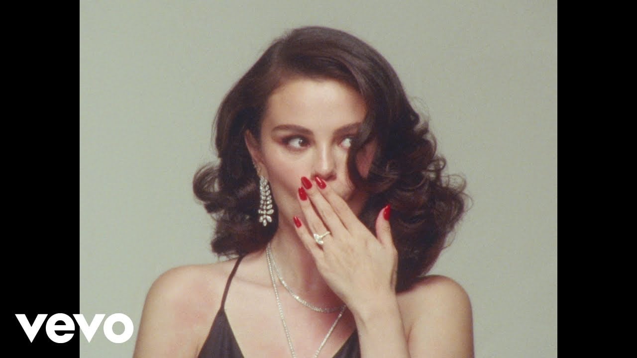 A woman with wavy dark hair, wearing a black top, dangling earrings, and necklaces, covers her mouth with her hand, showing red nails. The background is plain and light-colored. The Vevo logo is in the bottom left corner.