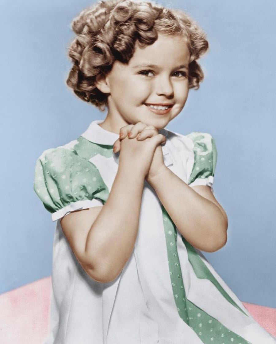 A young girl with curly hair smiles brightly, clasping her hands together. She wears a white dress with green polka-dotted puff sleeves and a matching sash, posed against a blue and pink background.