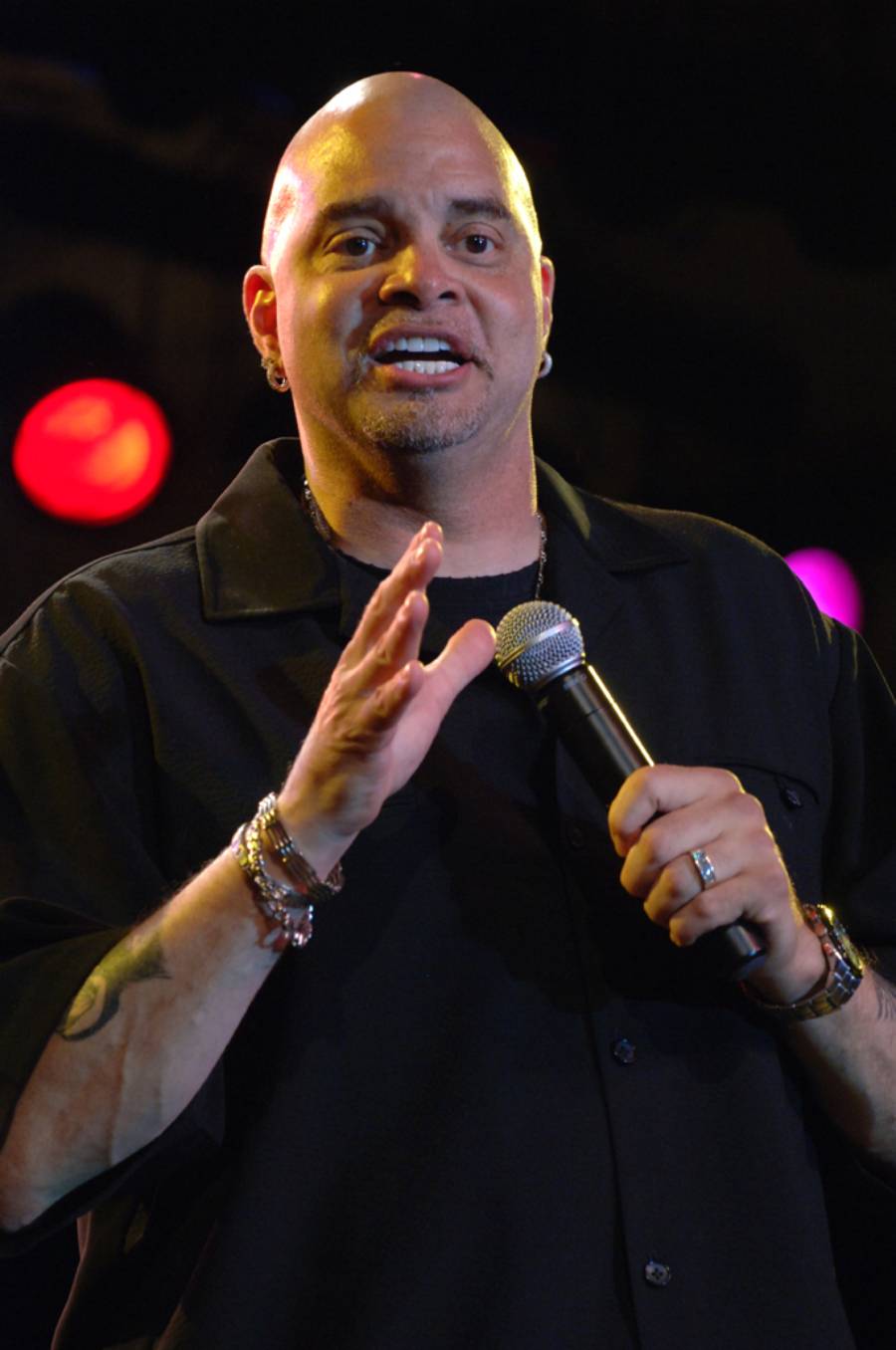 A bald man wearing a black shirt holds a microphone and gestures with one hand while performing on stage, with colorful lights blurred in the background.