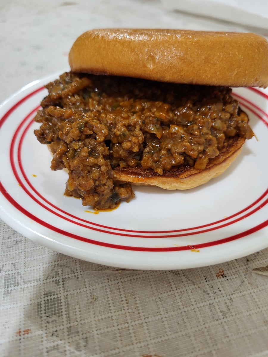 A sloppy joe sandwich with a generous serving of seasoned ground beef filling on a hamburger bun, served on a white plate with red stripes, set on a lace-patterned tablecloth.