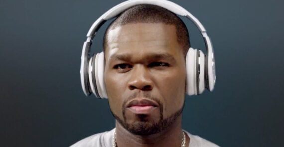 A man with a trimmed beard and mustache wears large white headphones over his ears, looking forward with a serious expression against a plain dark background.