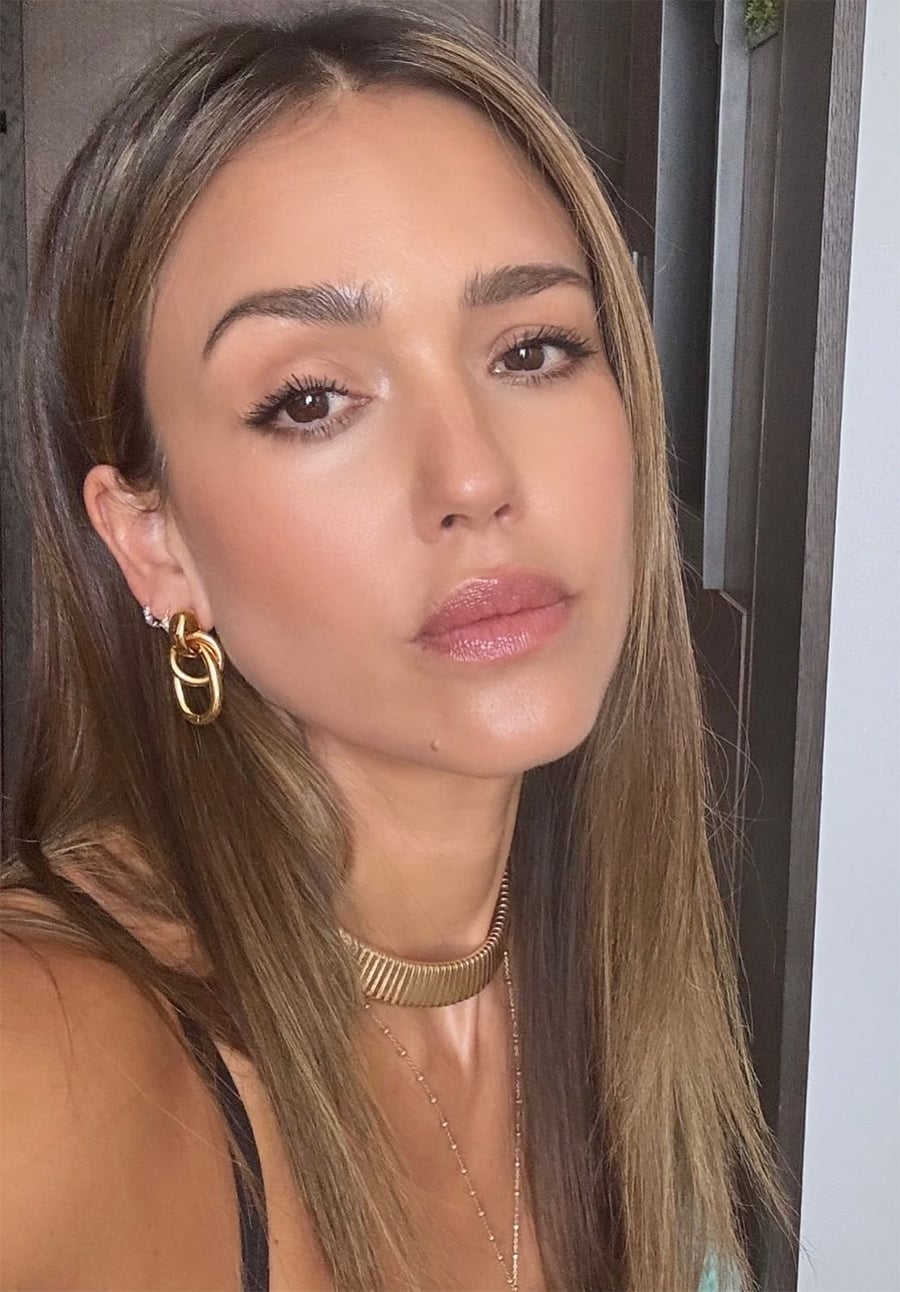 A woman with long, straight brown hair poses for a close-up selfie. She wears gold hoop earrings, a gold necklace, and natural makeup with glossy lips. The background appears to be indoors with neutral tones.