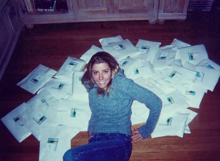 A woman in a gray sweater and jeans smiles while sitting on a wooden floor, surrounded by numerous white envelopes scattered around her.