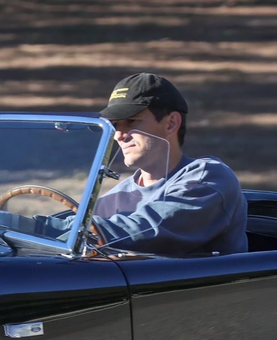 A person wearing a black cap and a blue sweatshirt drives a classic black convertible car with a wooden steering wheel on a sunny day.