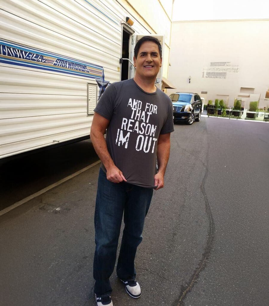 A man stands on a street next to a trailer, smiling at the camera. He is wearing a gray T-shirt that says “AND FOR THAT REASON I'M OUT” in large white letters, jeans, and sneakers. A black vehicle is parked in the background.
