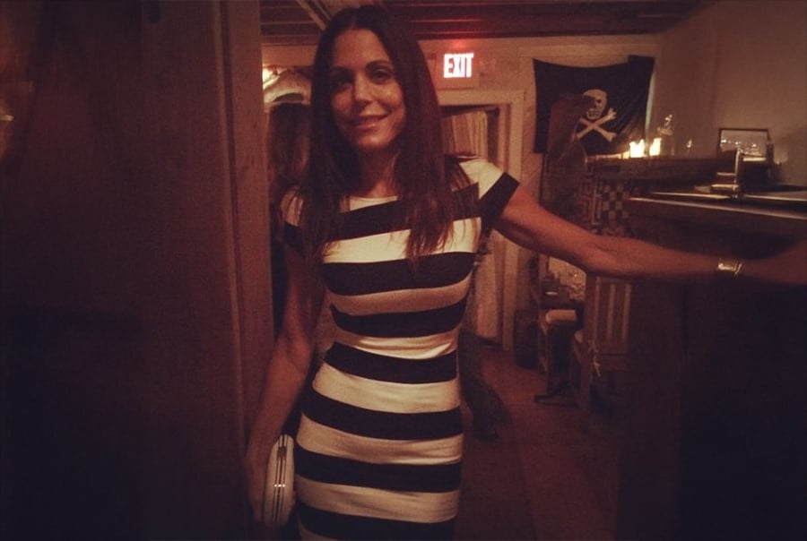 A woman in a black and white striped dress stands in a dimly lit room, smiling at the camera. There is a pirate flag and candles in the background, and an exit sign is visible above her.
