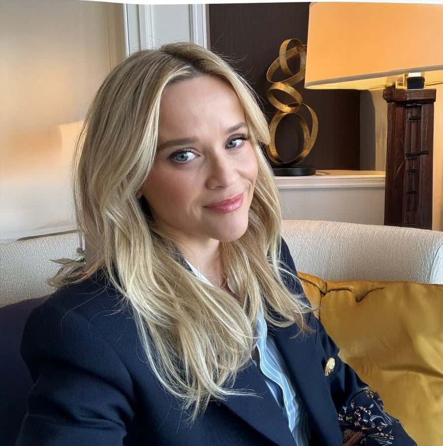 A woman with long blonde hair and a navy blazer sits on a cream couch, smiling softly at the camera. A yellow cushion and a modern lamp are visible in the background.