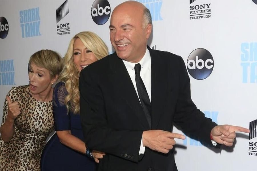 Three people dressed formally pose playfully on a Shark Tank-themed red carpet, pointing and smiling in front of a backdrop with ABC, Sony Pictures, and Shark Tank logos.