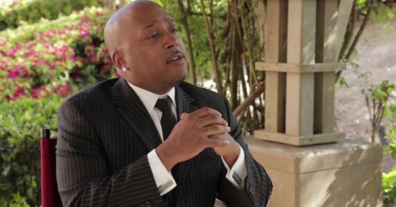 A man in a pinstripe suit and tie sits outdoors, clasping his hands while speaking. He is bald and wearing earrings, with greenery and flowers in the background.