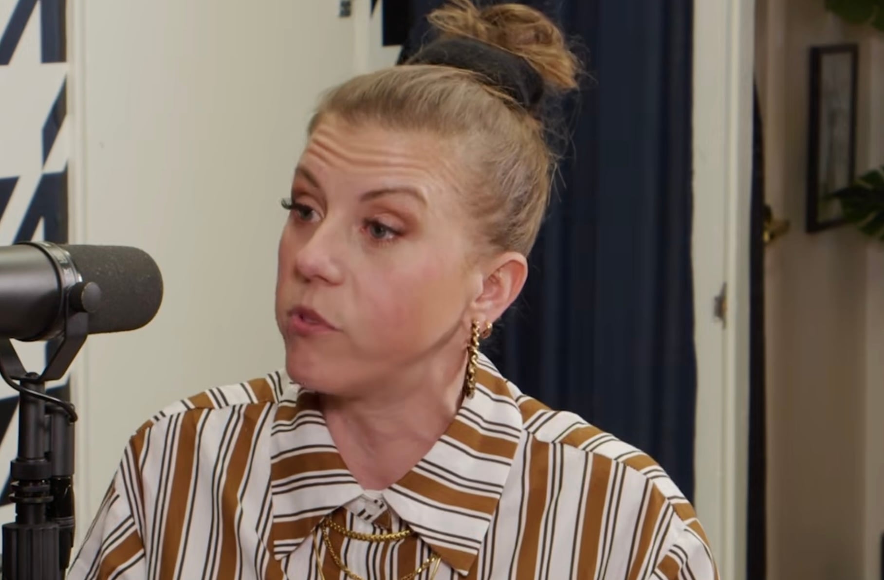 A woman with blonde hair in a bun, wearing gold hoop earrings and a brown and white striped shirt, speaks into a microphone during an indoor interview or podcast.