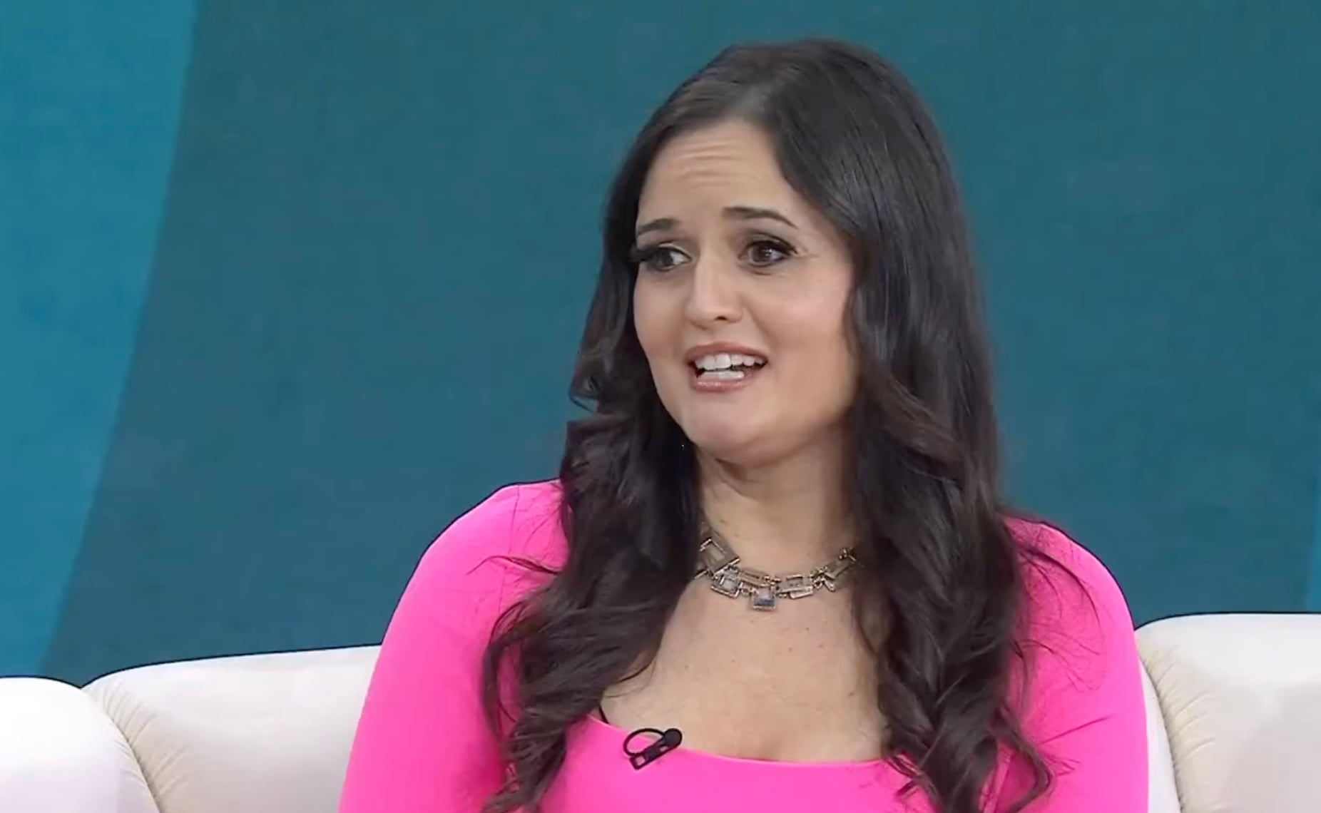 A woman with long dark hair wearing a bright pink top and a necklace sits on a white couch, speaking during what appears to be a television interview against a blue background.