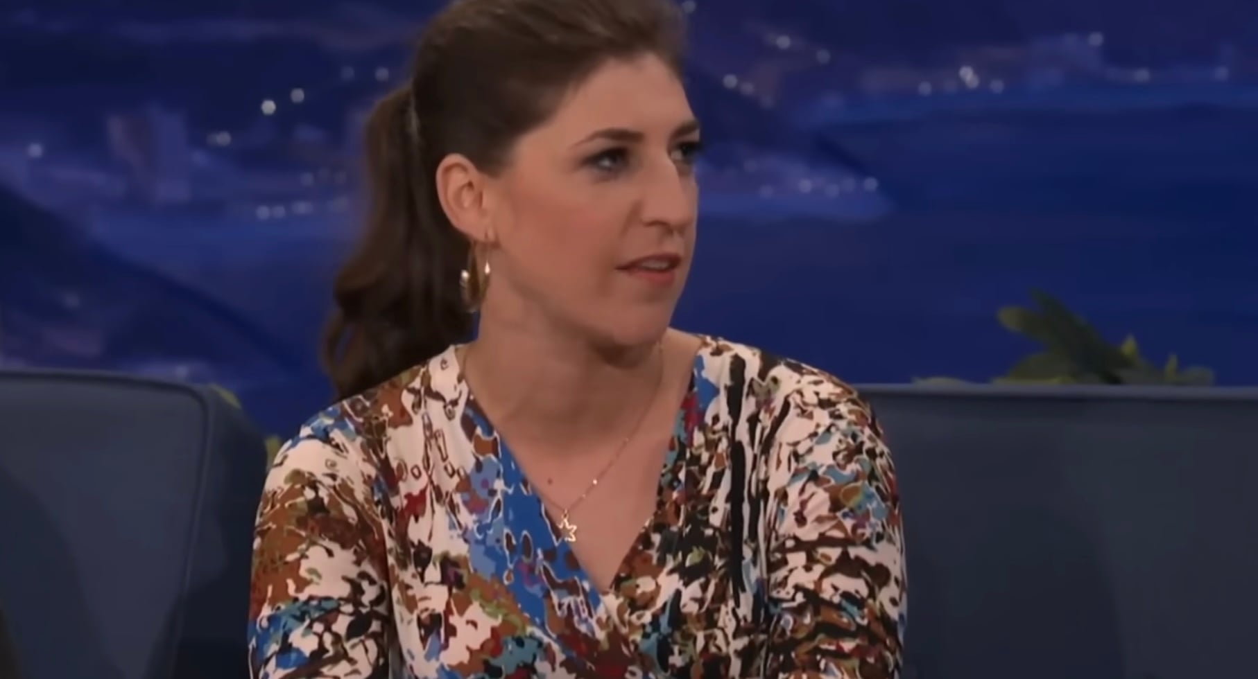 A woman with brown hair pulled back, wearing gold hoop earrings and a colorful patterned blouse, sits on a talk show set, looking to the side while speaking.