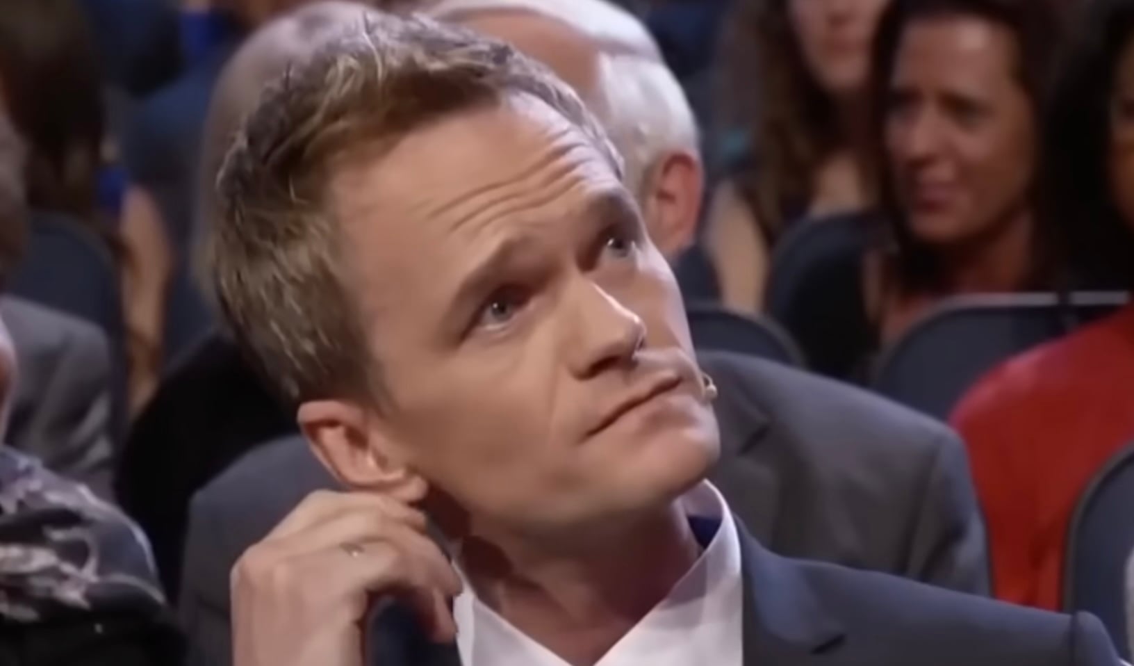 A man in a suit sits among a crowd, looking thoughtful while scratching the side of his head. The background is slightly out of focus, showing several other seated people.