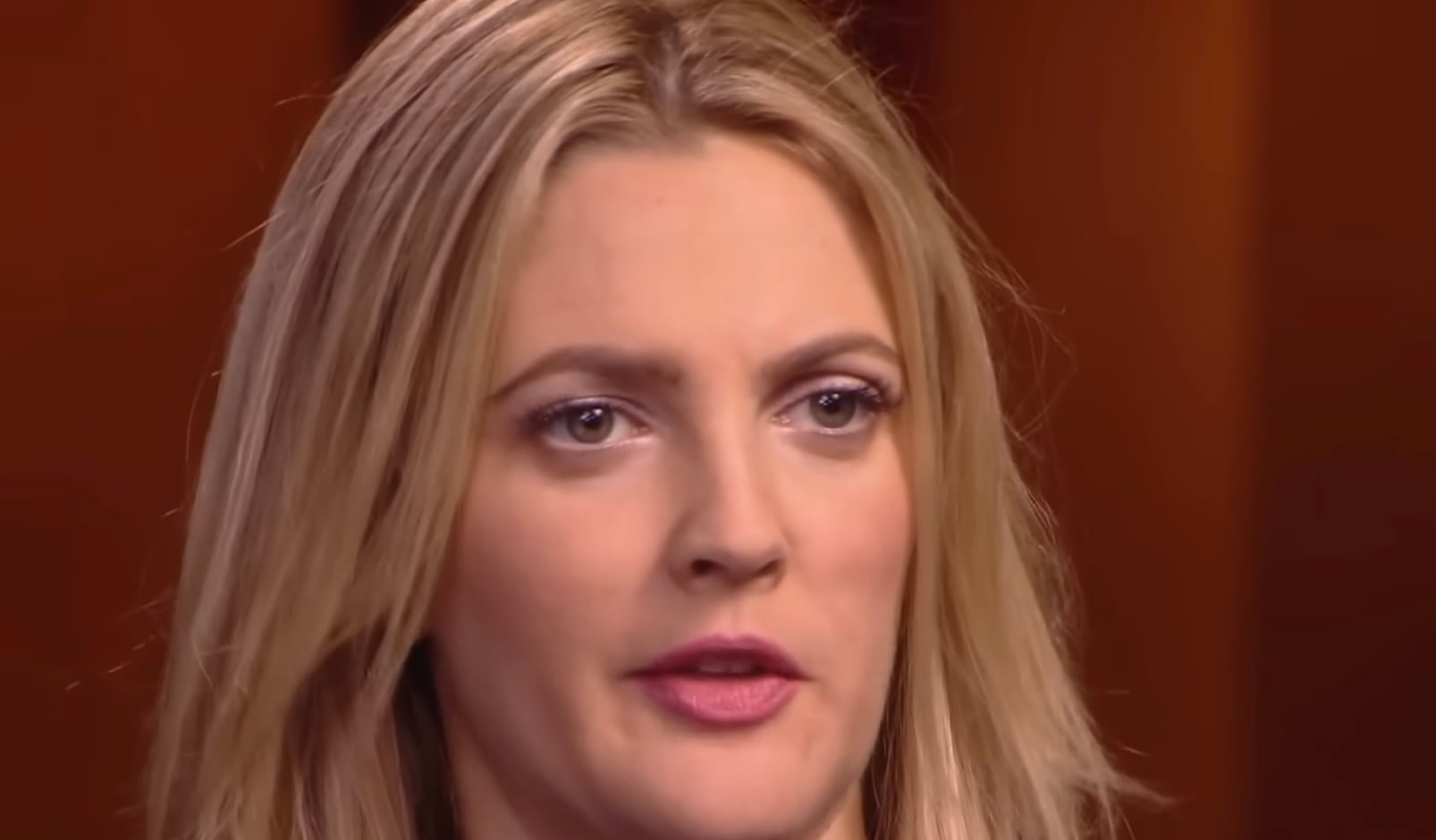 A woman with blonde hair and light makeup looks forward with a neutral expression against a softly blurred brown background.