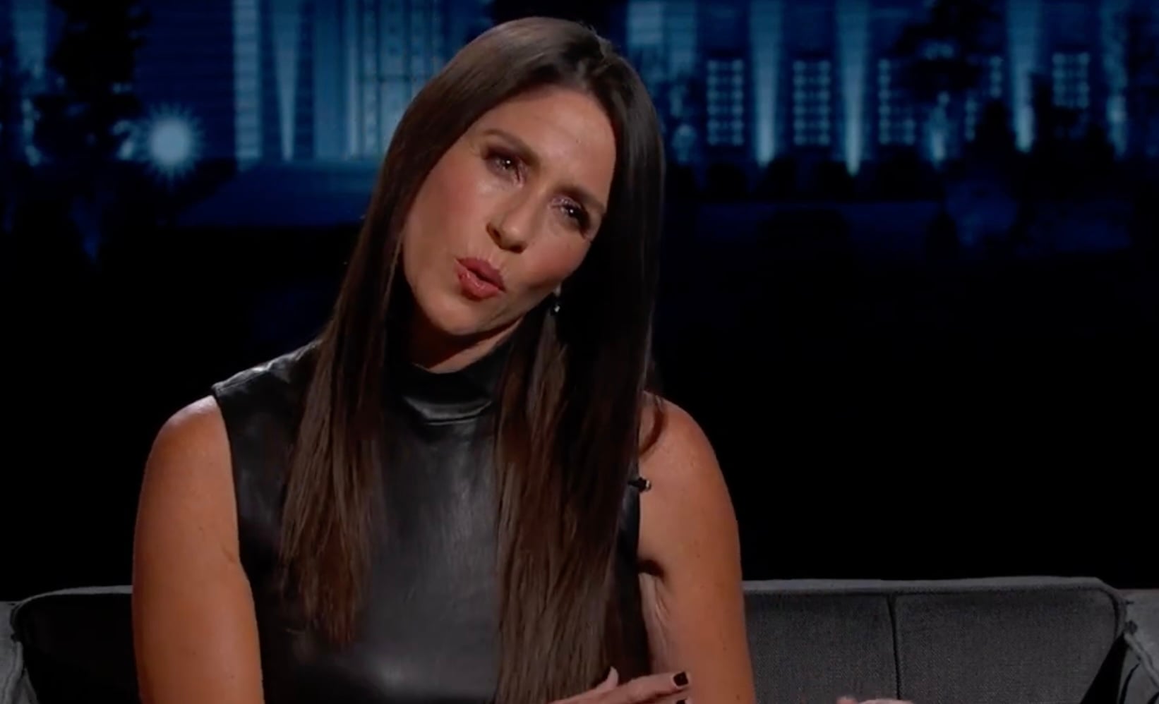 A woman with long brown hair, wearing a sleeveless black leather top, sits on a dark couch and tilts her head while pursing her lips, with a dark, blurred background behind her.