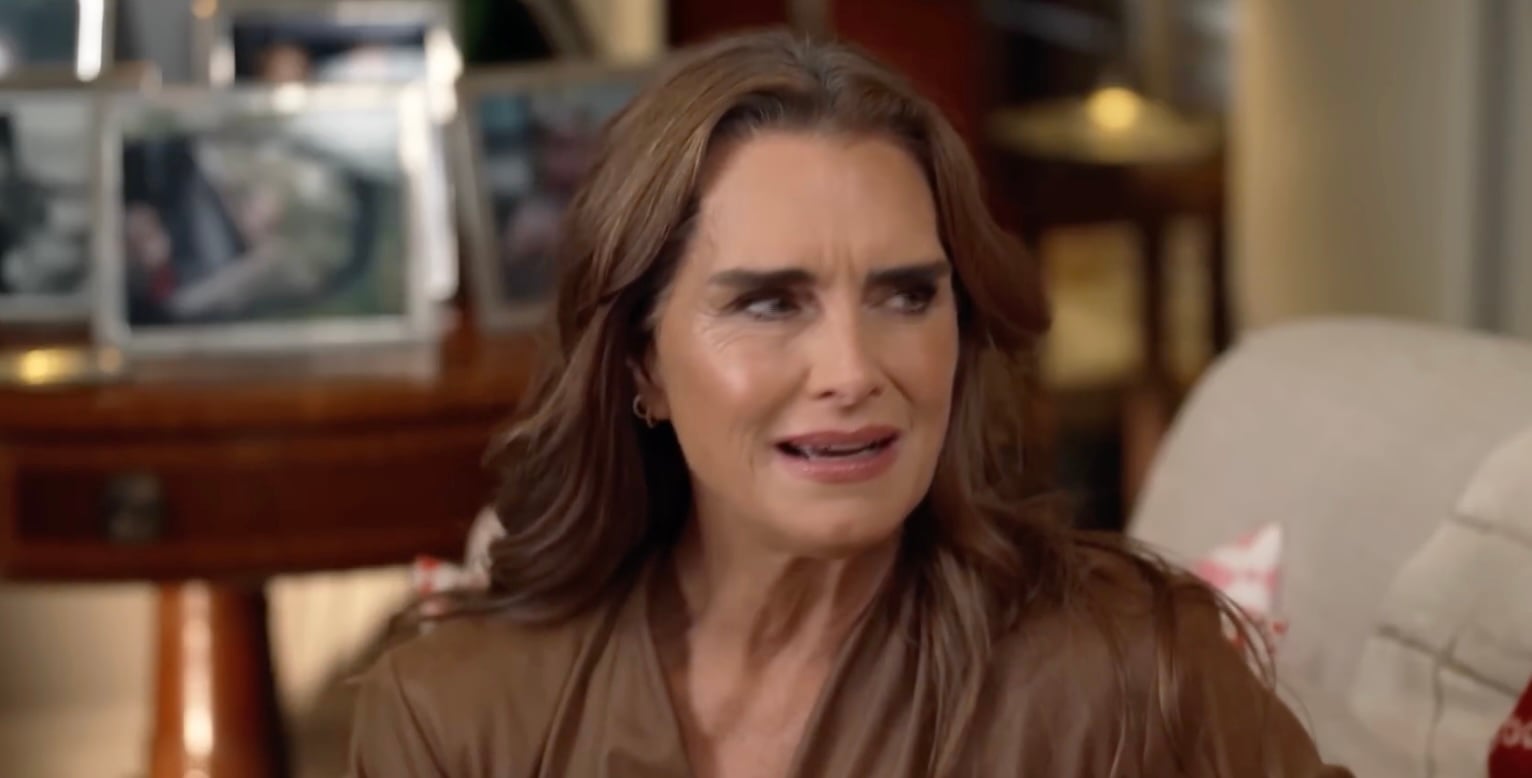 A woman with long brown hair and a tan leather top sits indoors, looking to the side with a confused or uncomfortable expression. Blurred photos and furniture are visible in the background.
