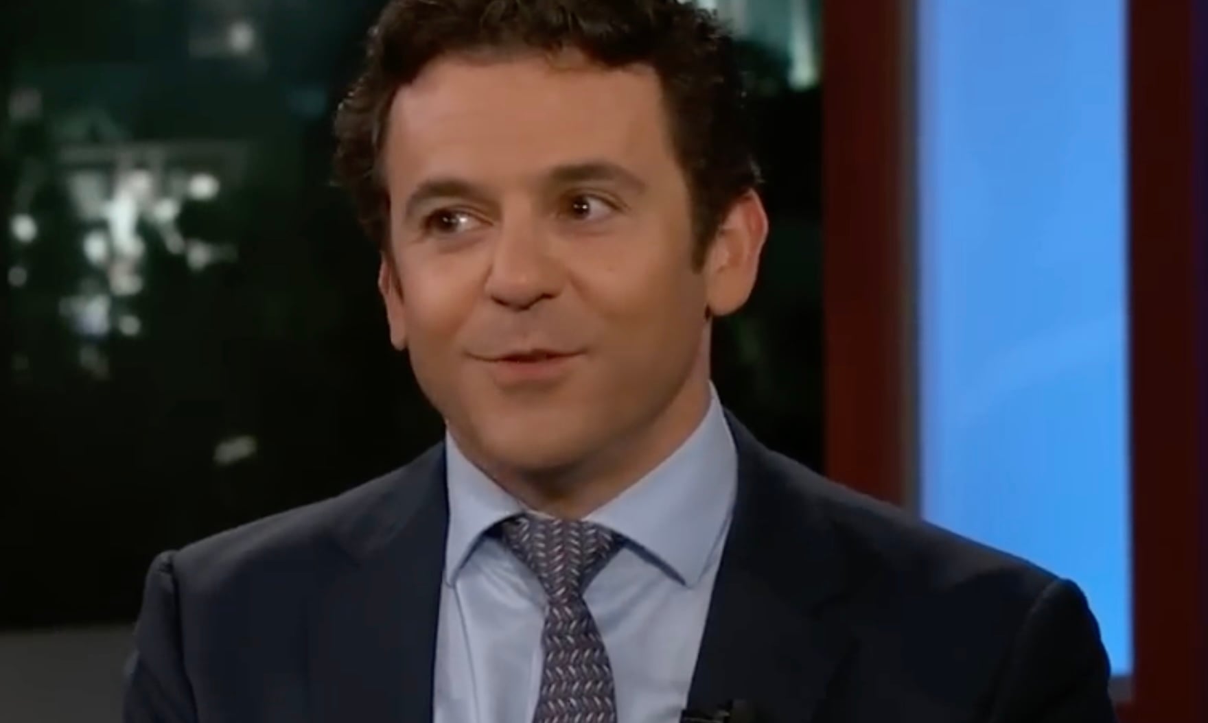 A man in a dark suit and patterned tie smiles while speaking during a television interview, with blurred studio lights and background visible.