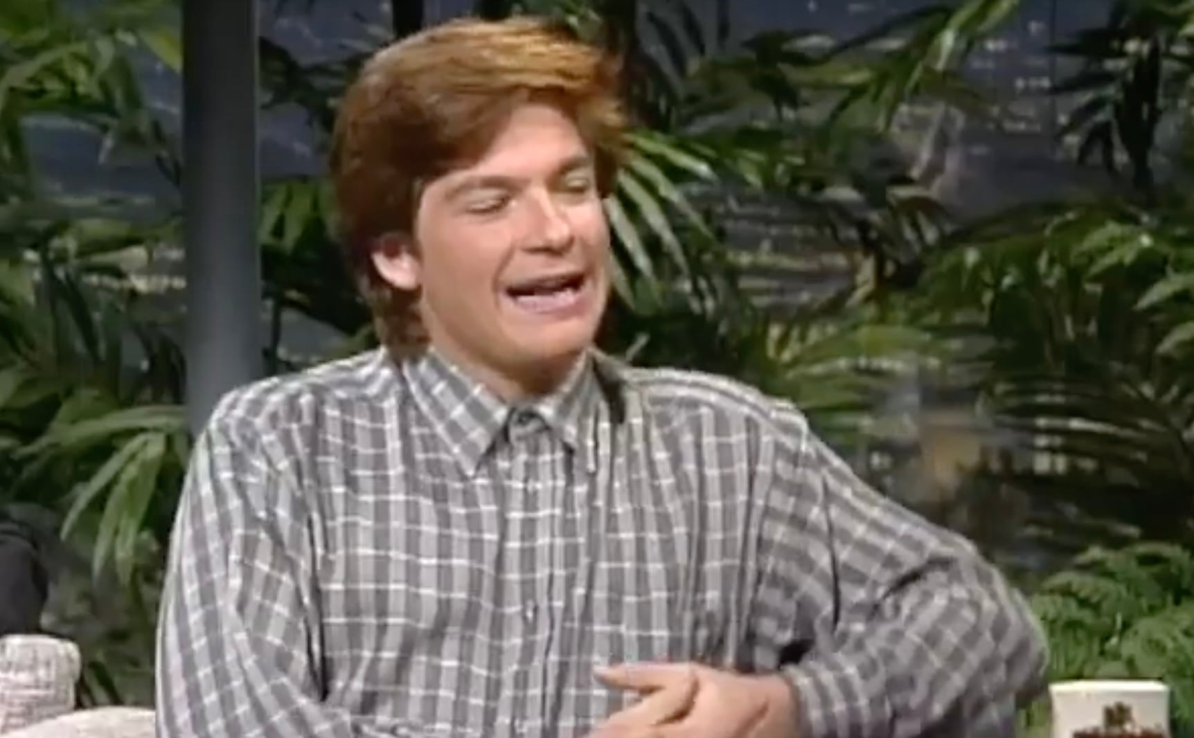 A person with light brown hair wearing a gray and white plaid button-up shirt sits indoors, smiling with eyes partially closed. There are large green plants and a blurred background behind them.