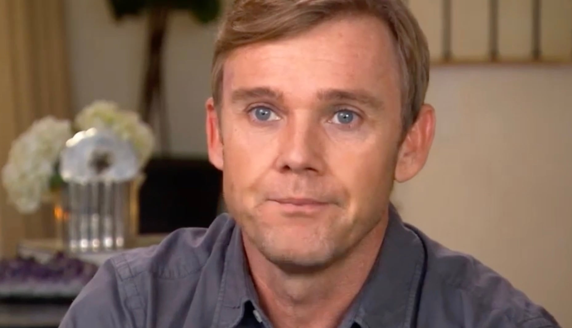 A man with light brown hair and blue eyes looks directly at the camera. He is wearing a gray shirt and is indoors with blurred flowers and decor in the background.