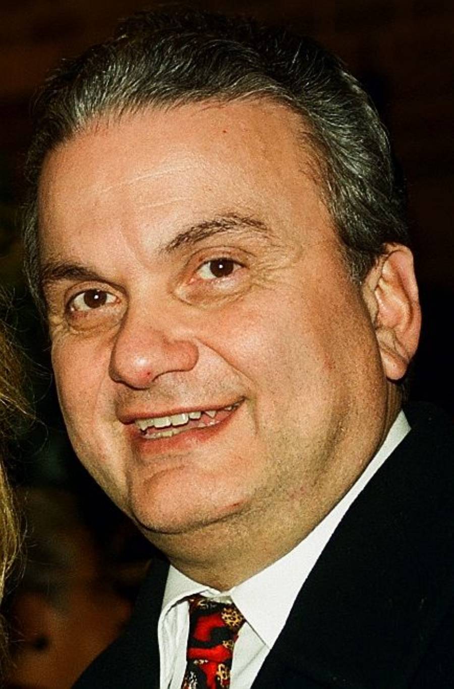A middle-aged man with gray hair smiles at the camera, wearing a dark jacket, white shirt, and a colorful tie. The background is out of focus.