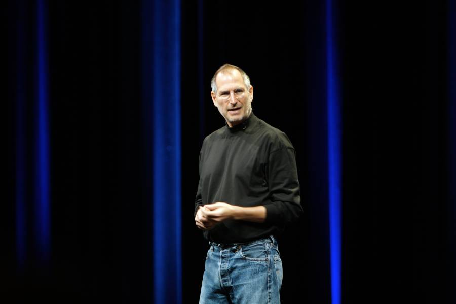 A man in a black turtleneck and blue jeans stands on stage in front of dark blue vertical lights, speaking and gesturing with his hands.
