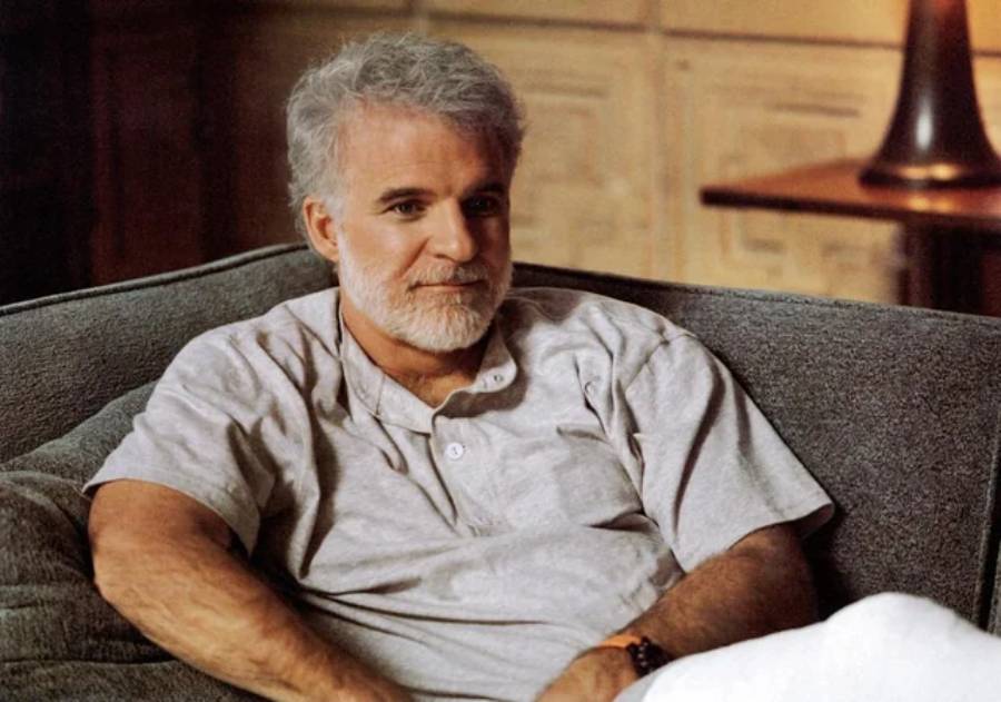 A man with gray hair and a beard sits on a gray couch, wearing a light gray short-sleeved shirt and white pants. He looks relaxed and is smiling slightly, with a soft, warm-toned background behind him.