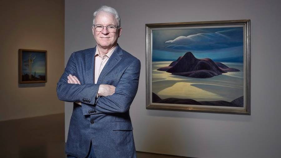 An older man with white hair and glasses, wearing a blue suit, stands with arms crossed in an art gallery. He is in front of a landscape painting featuring mountains and a dramatic sky. Another painting is visible in the background.