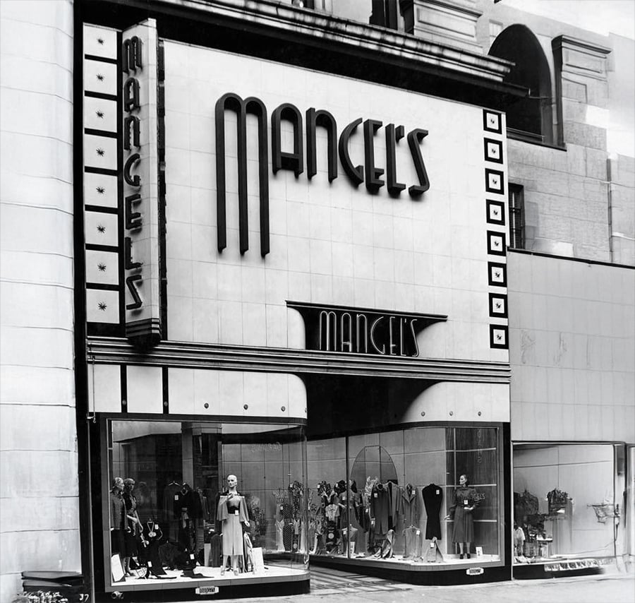 Black and white photo of Mangel’s, a vintage clothing store with mannequins dressed in women’s fashions displayed in large front windows. The storefront features bold, art deco style signage.