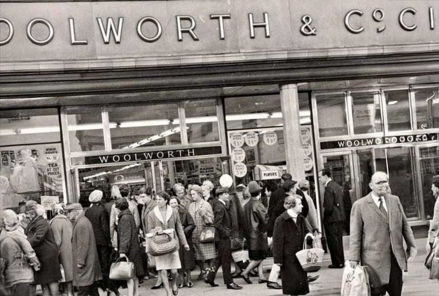 Crowds of people, mostly in coats and hats, gather outside a Woolworth store. The storefront has large windows and signs, and the scene appears to be from the mid-20th century.