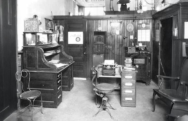 Black and white photo of an old-fashioned office with wooden furniture, a roll-top desk, typewriter, chairs, shelves with papers, and vintage office equipment against paneled walls.