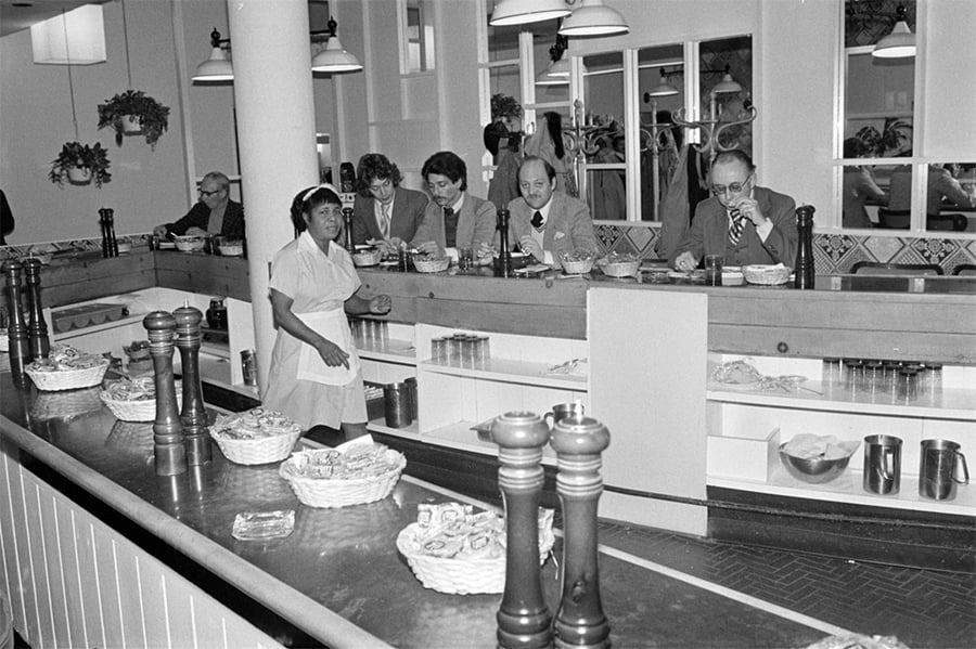 Black-and-white photo of a diner with a waitress walking behind the counter and several people seated, eating food. Decorative plants hang on the walls and baskets with food are placed on the counter.