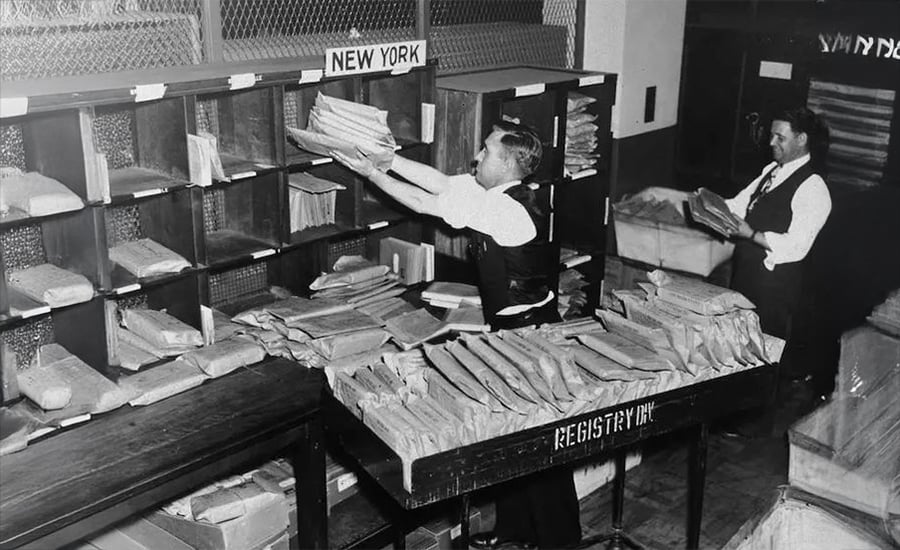 Two men sort and organize large stacks of mail on shelves labeled "New York" in a postal registry office. The workspace is filled with packages and envelopes, and both men are wearing dress shirts and ties.