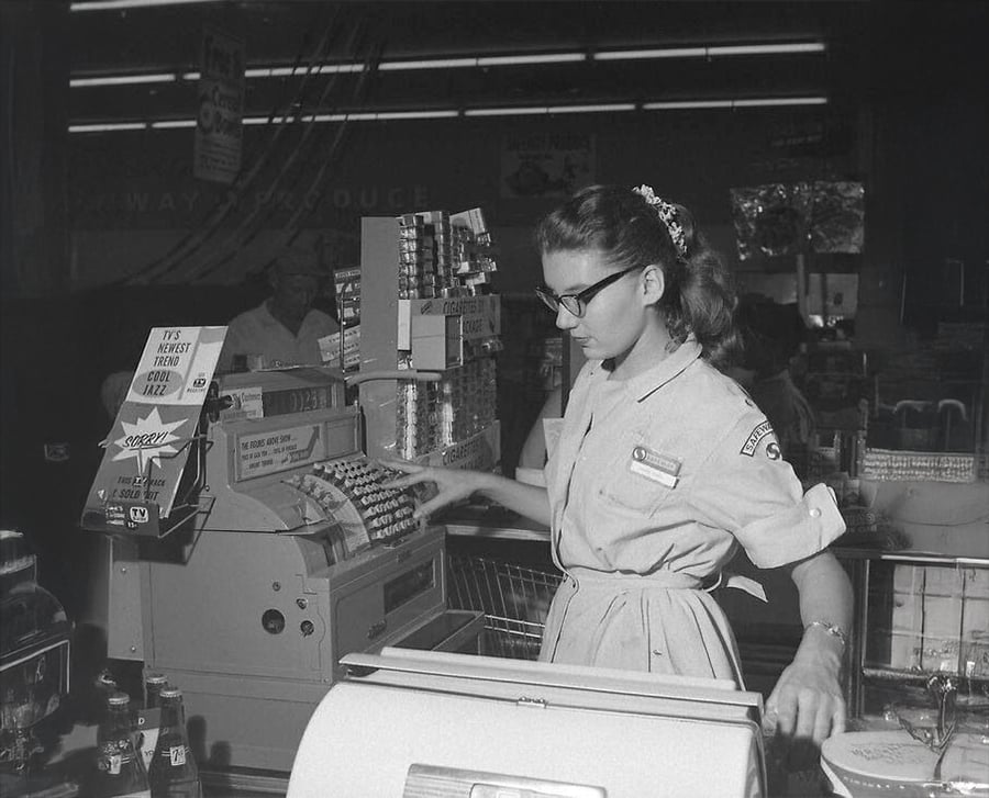 A woman in a uniform dress and glasses operates a vintage cash register at a grocery store checkout counter, surrounded by products and store displays, in a black-and-white photo.