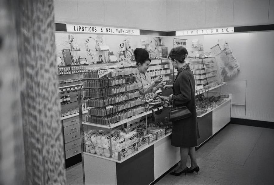 Two women stand at a cosmetics counter in a store, one behind the counter assisting the other. Shelves are lined with lipstick, nail varnish, and various beauty products. The scene appears to be from the mid-20th century.