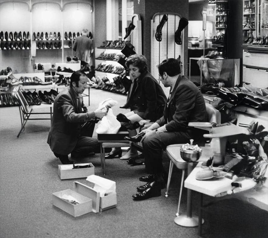 A shoe salesman assists a woman trying on shoes while a man sits beside her in a 1960s shoe store filled with displays of various footwear.