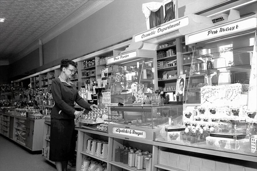 A woman in a dark dress stands at a cosmetics counter inside a vintage pharmacy or department store, surrounded by perfume bottles, makeup products, and shelves stocked with various goods.