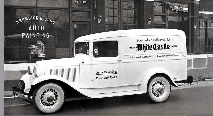 A vintage white delivery van parked in front of an auto painting shop. The van advertises "White Castle" buns and "Johns Bake Shop" with a phone number on the door. The street scene appears from the early 20th century.