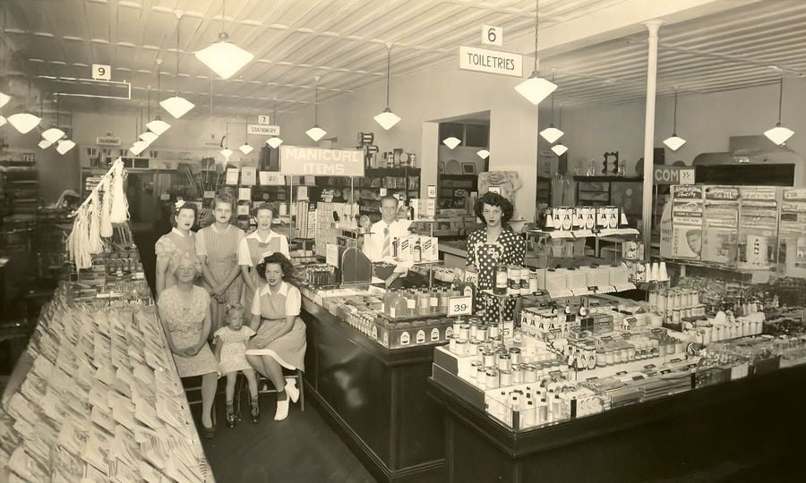 Black-and-white photo of a vintage drugstore with shelves of toiletries and personal care items. Several women, men, and children pose among the displays. Signs read “Manicure Items” and “Toiletries.” Bright ceiling lights hang above.