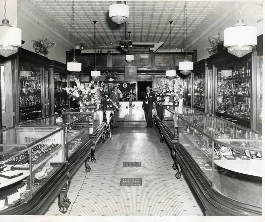 Black-and-white photo of a vintage jewelry store interior with glass display cases on both sides, floral arrangements, and two people standing at the back of the store near the counter.