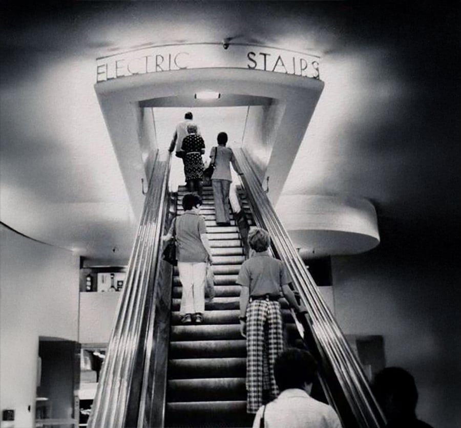 Black-and-white photo of people riding an escalator under a sign reading "Electric Stairs." The scene appears to be in a department store or public building, with vintage 20th-century clothing styles.
