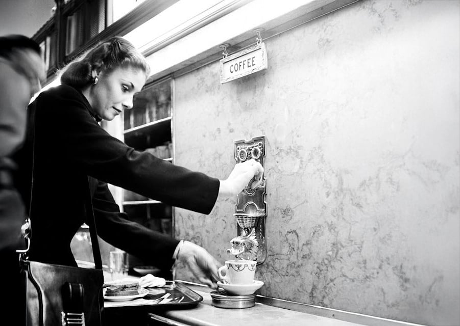 A woman in a coat dispenses coffee from a wall-mounted coffee machine into a cup at a counter. A small sign above the machine reads "COFFEE." She carries a purse and has plates with food nearby. The scene appears vintage.